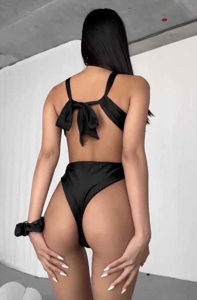 Back view of a black cutout monokini with crisscross straps and bow detail, worn by a woman standing in a minimalist indoor setting.